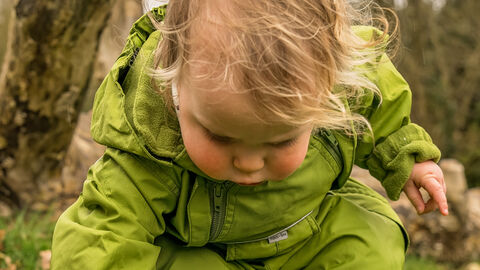 toddler wearing green puddlesuit and blue wellies reaching down to pick up stick
