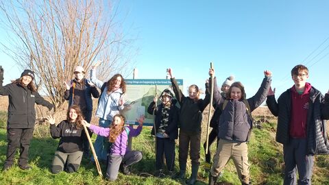 Diverse group of young people with shovels stand outside a nature reseve, smiling at the camera