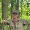 A man smiles while he leans against a tree in a wooded area